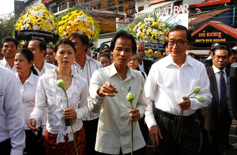 Chea Mony, wearing a white shirt and holding a green flower, walks with others also wearing white shirts and holding flowers. Bouquets of yellow flowers and a video rental store are seen in the background.